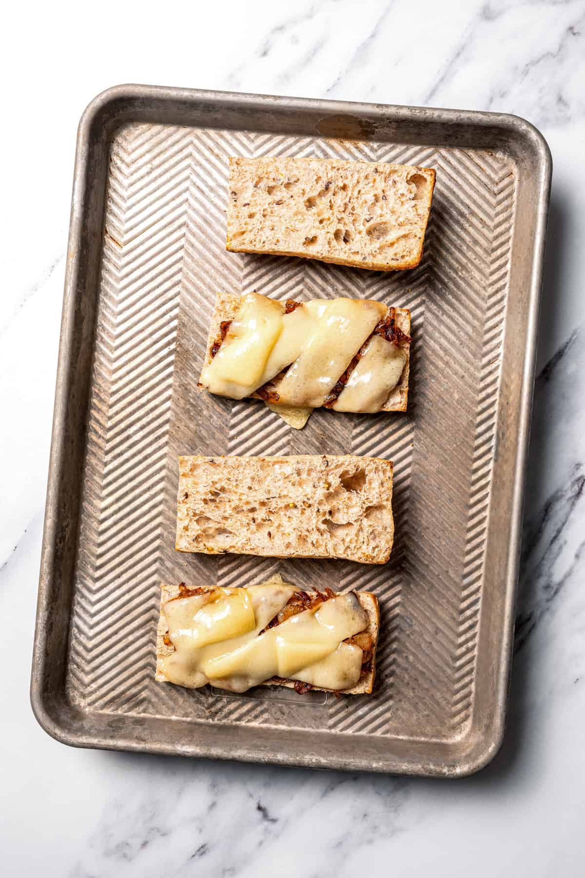 Overhead view of ciabatta roll halves topped with melted cheese on a baking sheet.