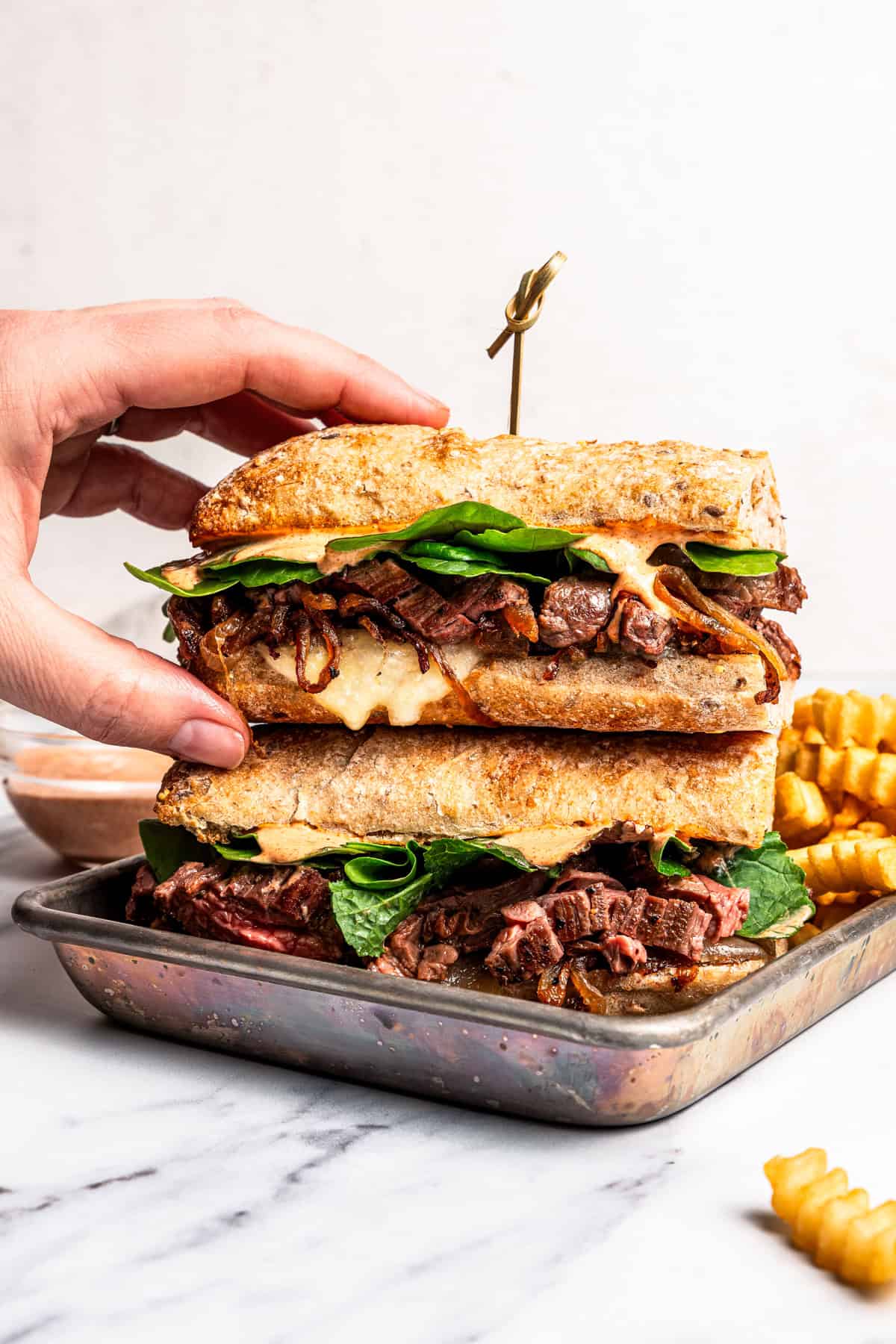 A hand reaching for one half of a steak sandwich stacked on a tray.