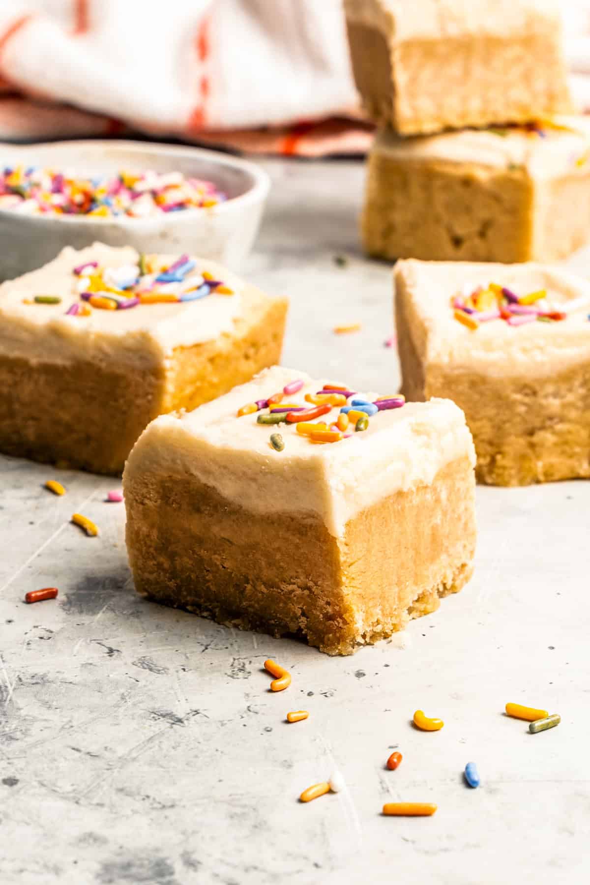 Mixing cookie butter into the dough makes the best sugar cookie bars ever! These bars are soft and chewy, topped with creamy frosting. Sugar cookie bars topped with frosting and sprinkles on a countertop, with a bowl of sprinkles in the background.