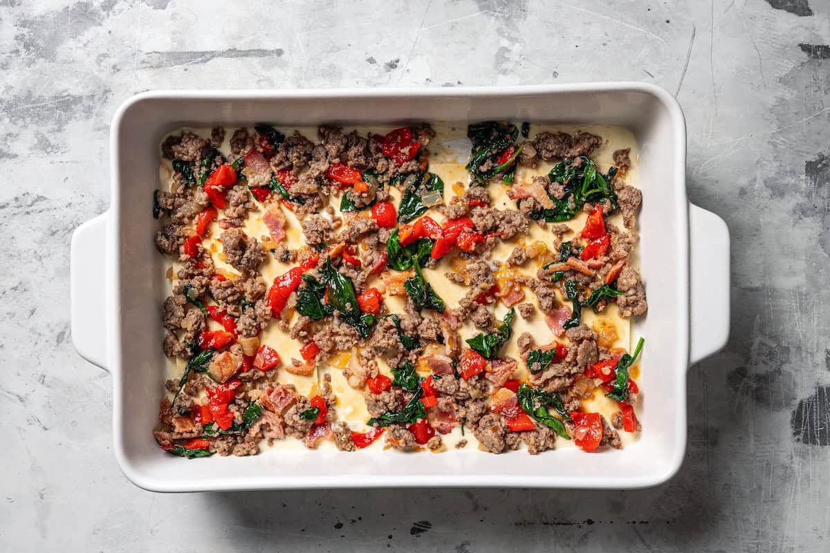 Breakfast sausage, bacon, and veggies layered over lasagna sheets and cheese sauce in a baking dish.