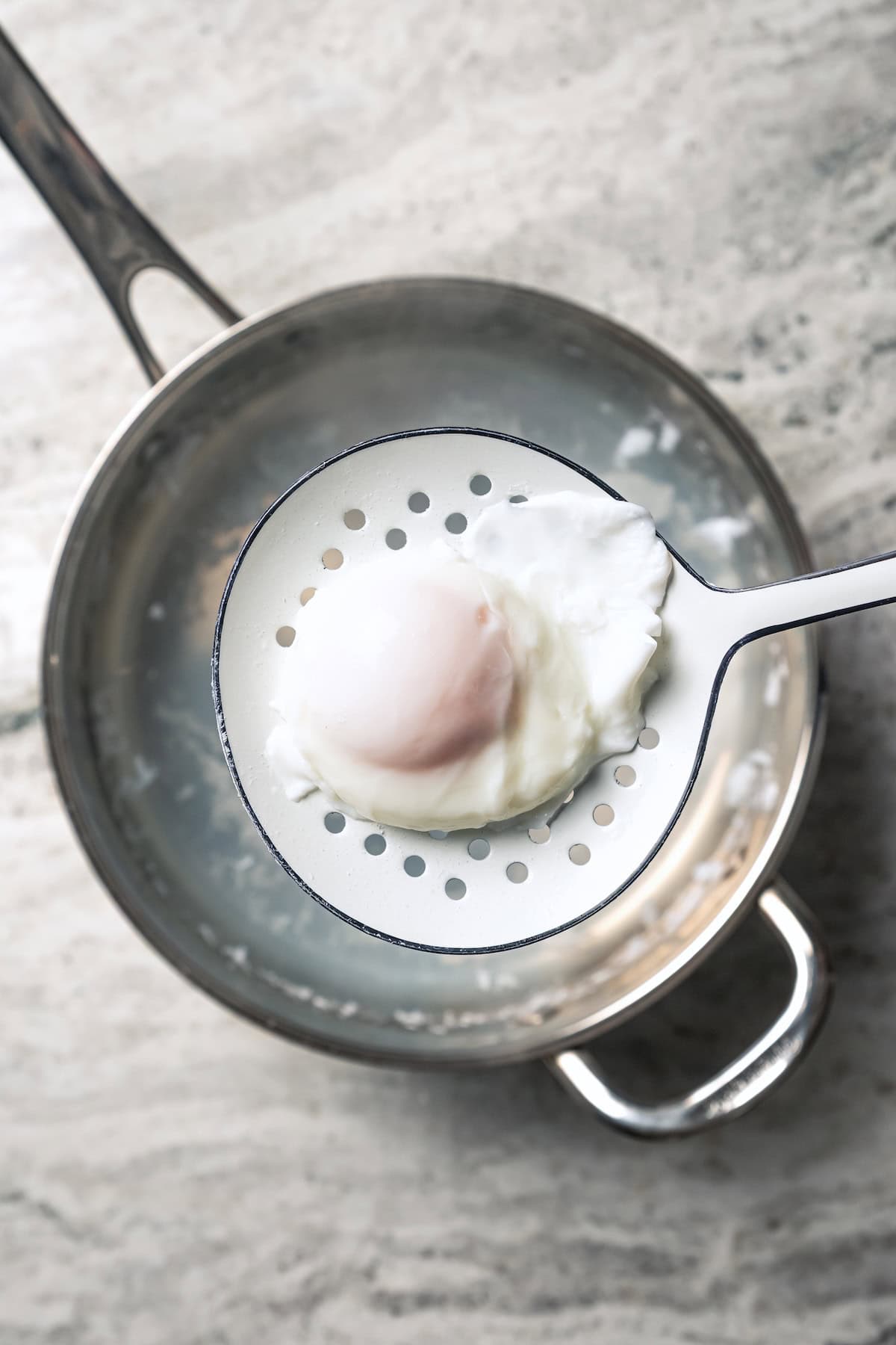 A poached egg being lifted from a pot of simmering water with a slotted spoon.
