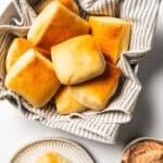 Texas Roadhouse rolls in a bowl lined with a dishcloth next to a sliced roll topped with cinnamon butter on a plate.