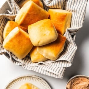 Texas Roadhouse rolls in a bowl lined with a dishcloth next to a sliced roll topped with cinnamon butter on a plate.