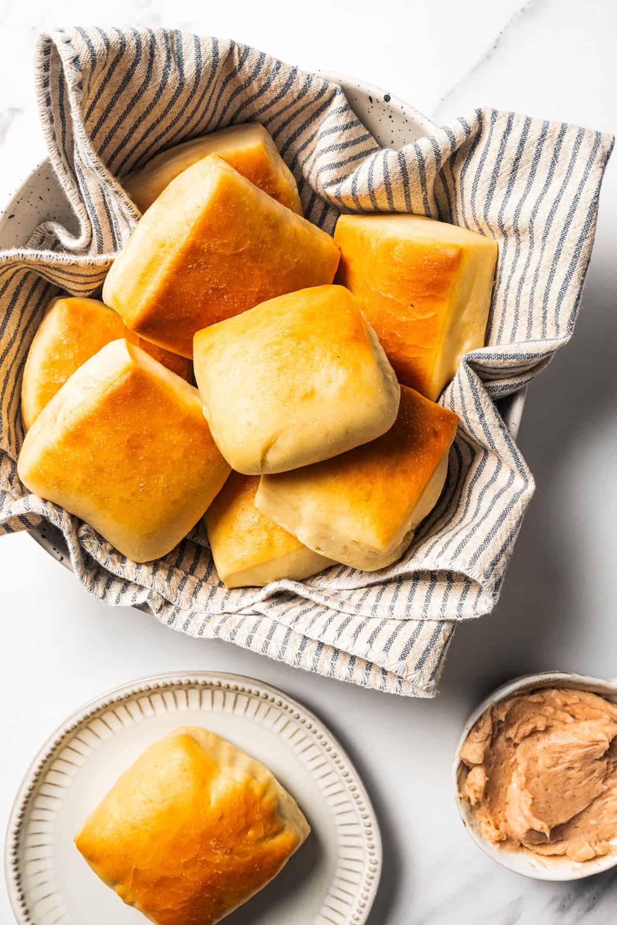 Texas Roadhouse rolls in a bowl lined with a dishcloth next to a sliced roll topped with cinnamon butter on a plate.