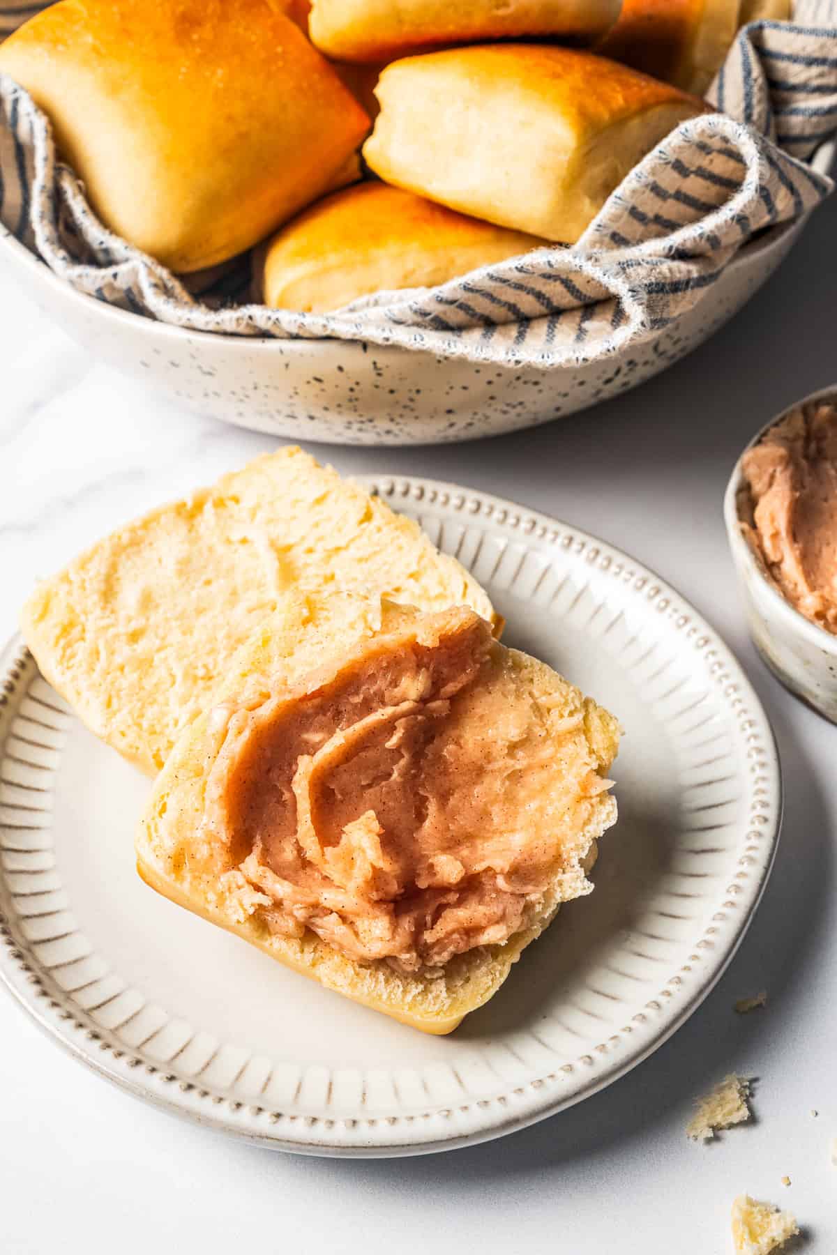 A Texas Roadhouse roll topped with cinnamon butter on a plate, with more rolls in a bowl in the background.