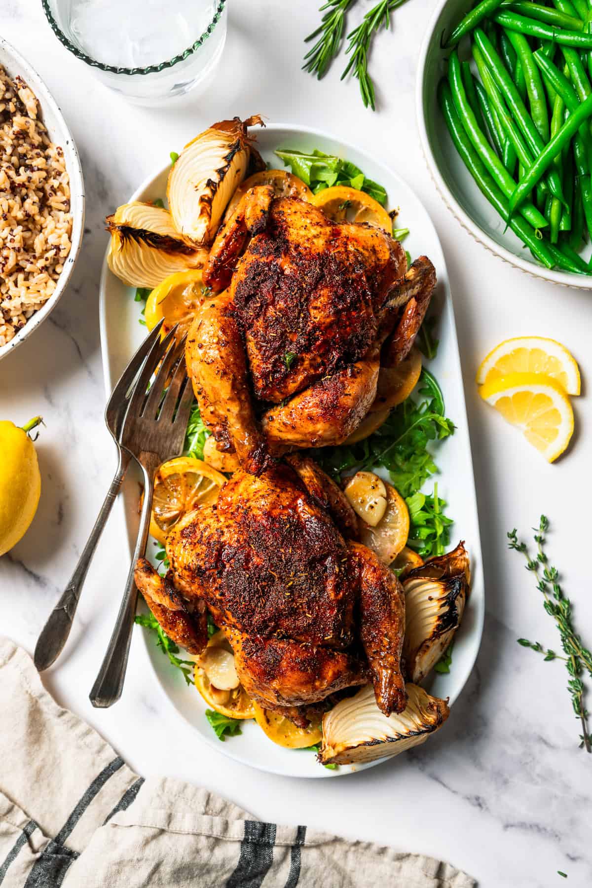 This easy Cornish hen recipe is perfectly juicy and golden-brown, rubbed with savory spices and oven-roasted over a bed of lemon and herbs. Two Cornish hens on a platter, with a bowl of rice set on one side of the platter, and a bowl of cooked green beans in another bowl.