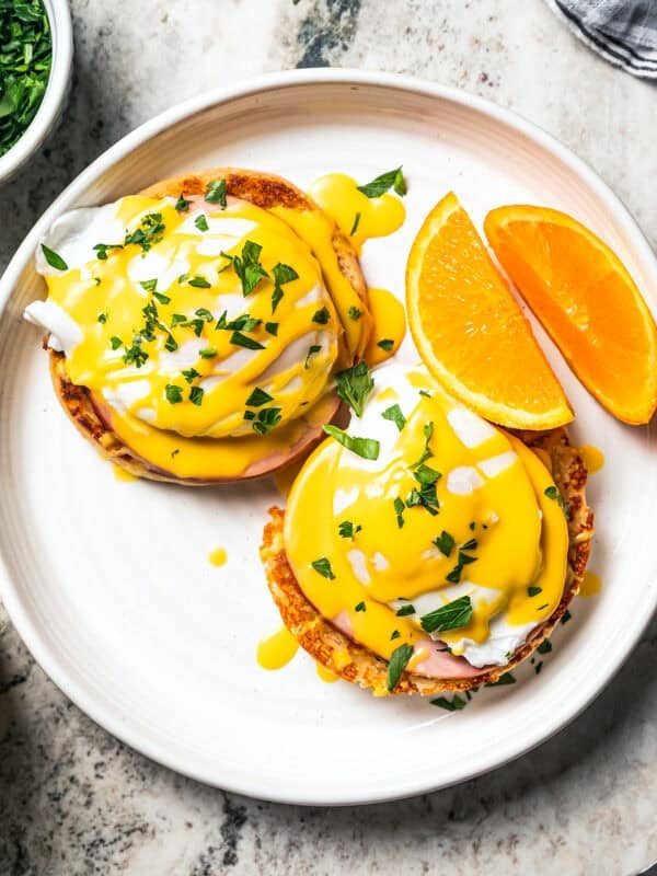 Eggs Benedict served with orange wedges on a plate next to a jar of Hollandaise sauce.