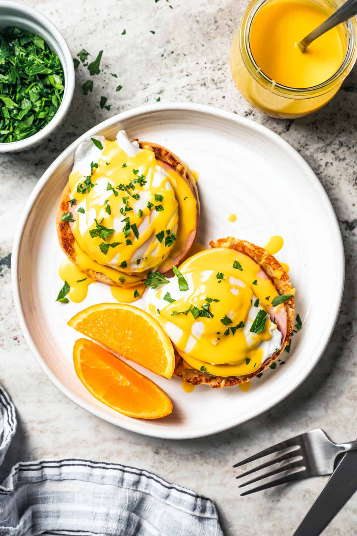 Eggs Benedict served with orange wedges on a plate next to a jar of Hollandaise sauce.