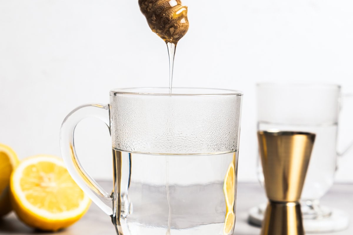 This classic hot toddy recipe is a soothing blend of your favorite whiskey, a drop of honey, and a squeeze of lemon juice with hot water. Adding honey to hot water in a glass mug to make hot toddies.