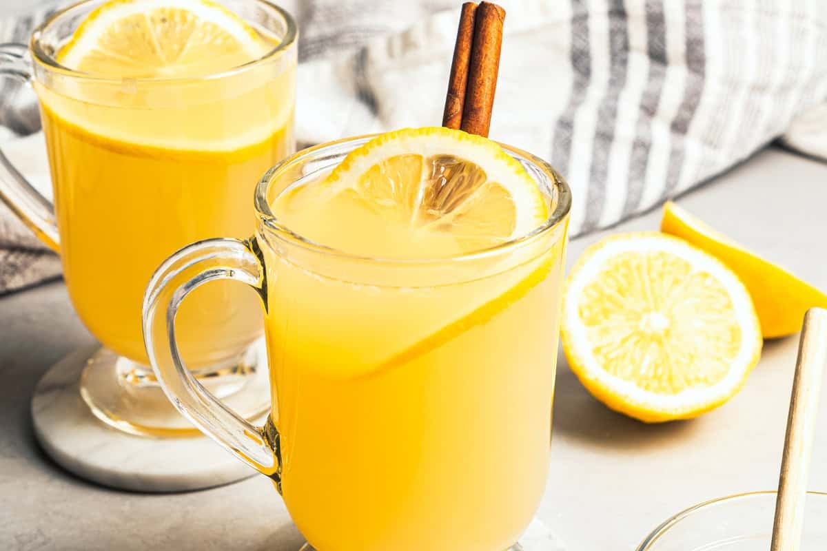 This classic hot toddy recipe is a soothing blend of your favorite whiskey, a drop of honey, and a squeeze of lemon juice with hot water. A hot toddy in a glass mug garnished with a fresh lemon slice and a cinnamon stick, with a second hot toddy in the background.