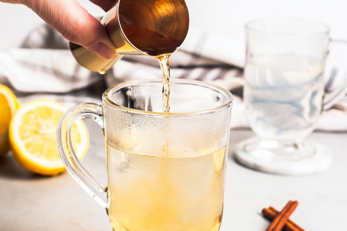 This classic hot toddy recipe is a soothing blend of your favorite whiskey, a drop of honey, and a squeeze of lemon juice with hot water. Pouring bourbon into a glass mug with hot water and honey.