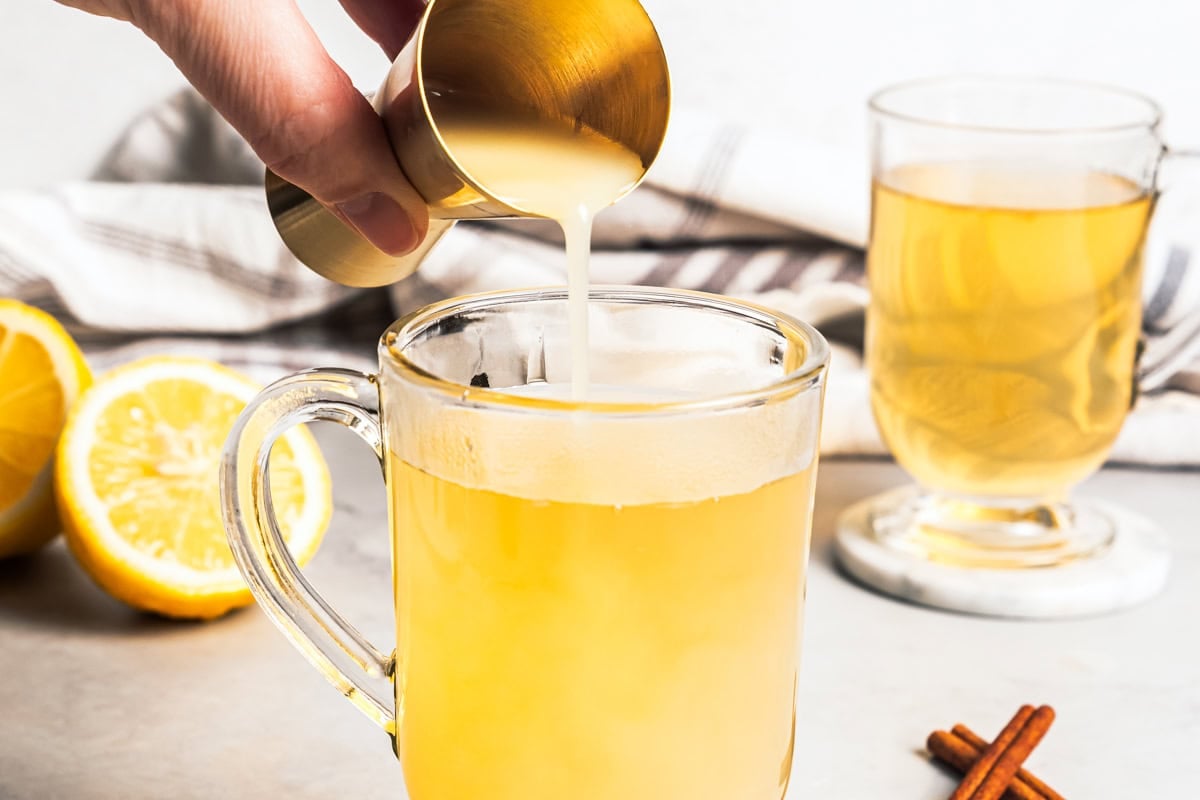 Pouring lemon juice into a glass mug with hot water, honey, and bourbon.