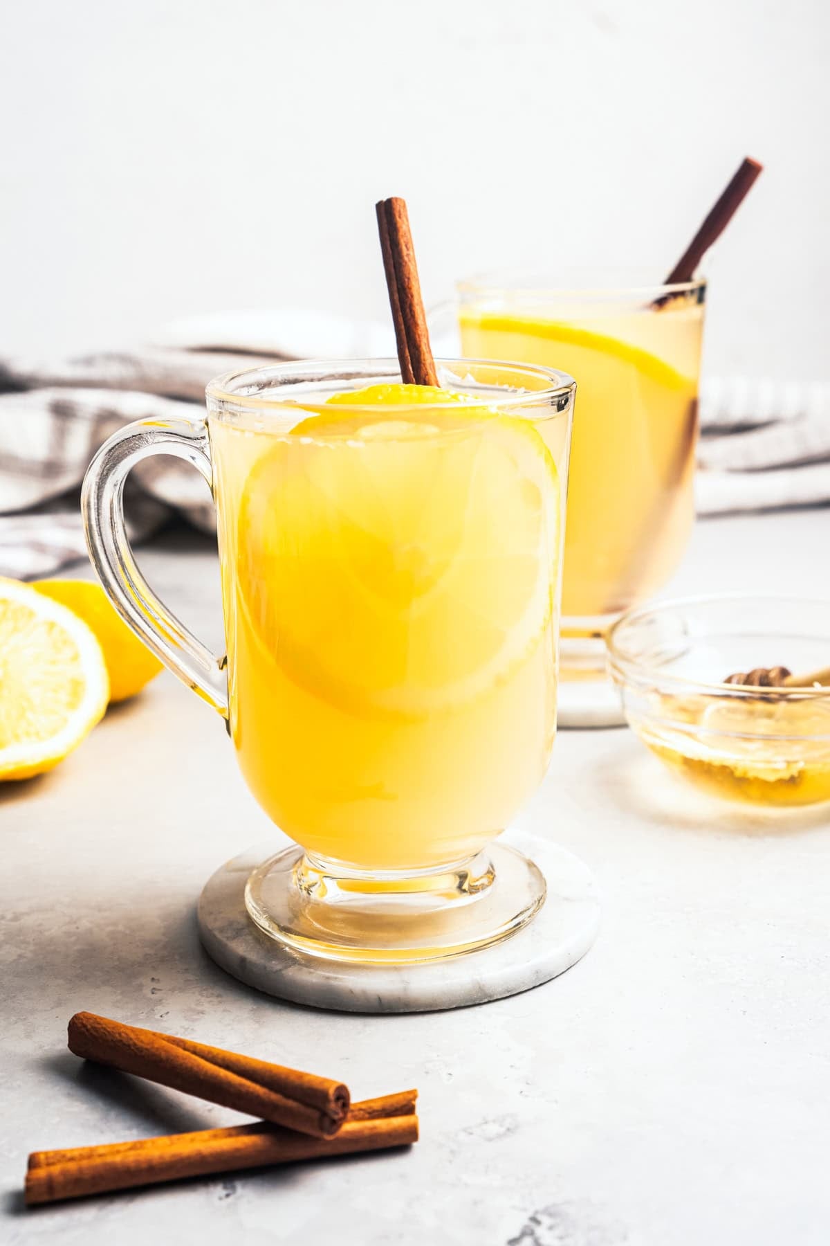 This classic hot toddy recipe is a soothing blend of your favorite whiskey, a drop of honey, and a squeeze of lemon juice with hot water. A hot toddy in two glass mugs garnished with a fresh lemon slice and a cinnamon stick.