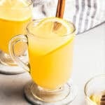 A hot toddy in a glass mug garnished with a fresh lemon slice and a cinnamon stick, with a second hot toddy in the background.