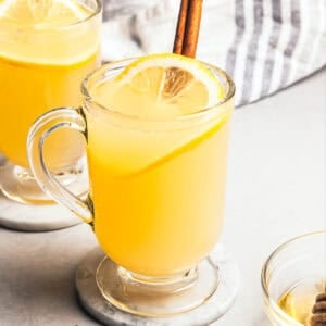 A hot toddy in a glass mug garnished with a fresh lemon slice and a cinnamon stick, with a second hot toddy in the background.