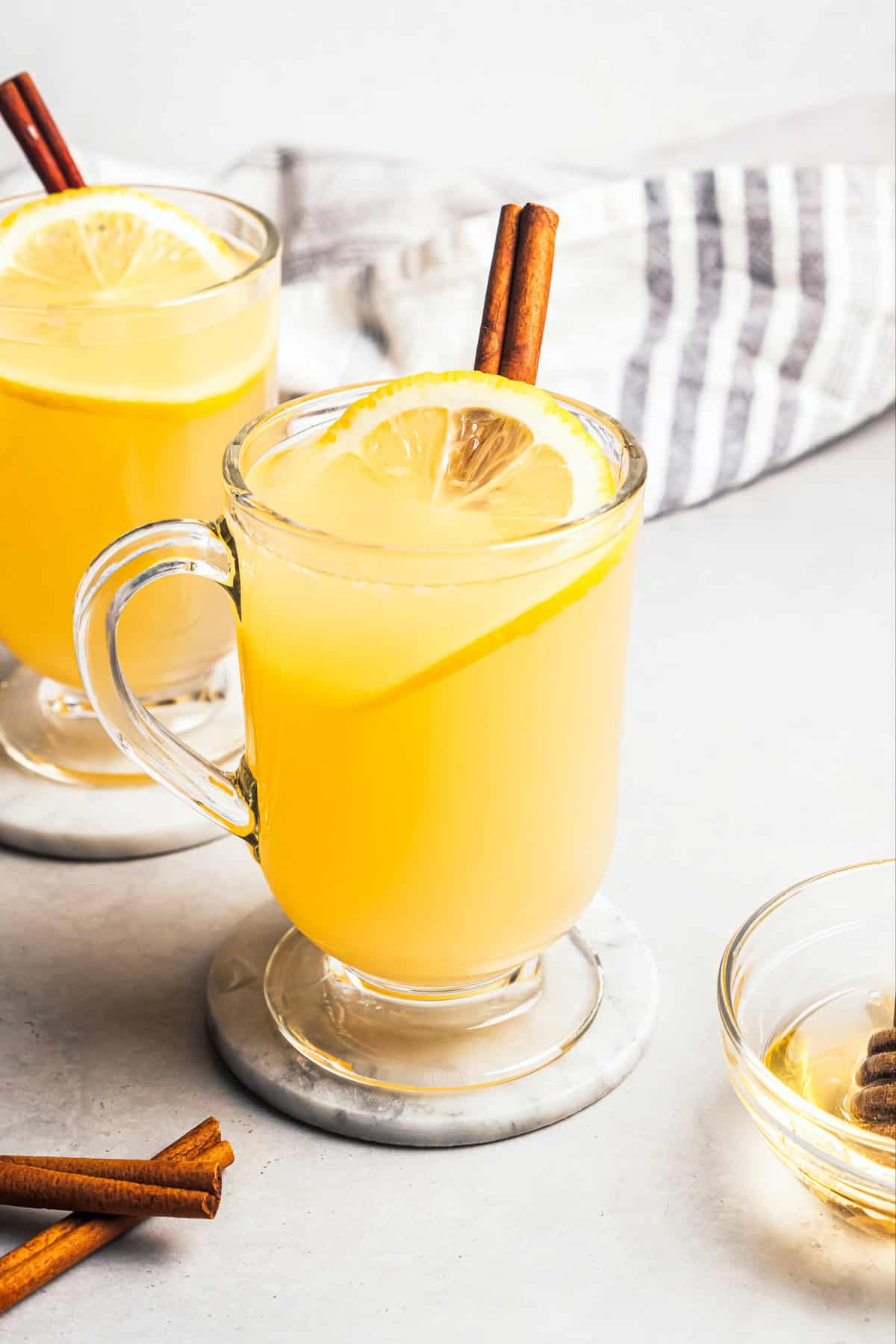 This classic hot toddy recipe is a soothing blend of your favorite whiskey, a drop of honey, and a squeeze of lemon juice with hot water. A hot toddy in a glass mug garnished with a fresh lemon slice and a cinnamon stick, with a second hot toddy in the background.