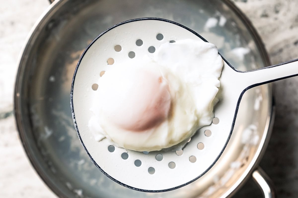 A poached egg is being lifted from a pot of simmering water with a slotted spoon.
