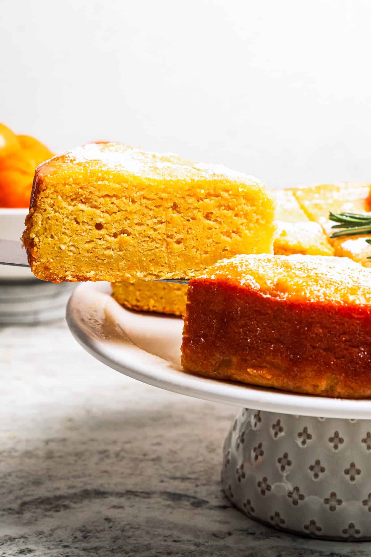 This orange cake recipe uses whole oranges, peels and all! It's moist and full of sweet citrus flavor, drenched in orange glaze. A slice of orange cake being lifted from the rest of the cake on a cake stand.