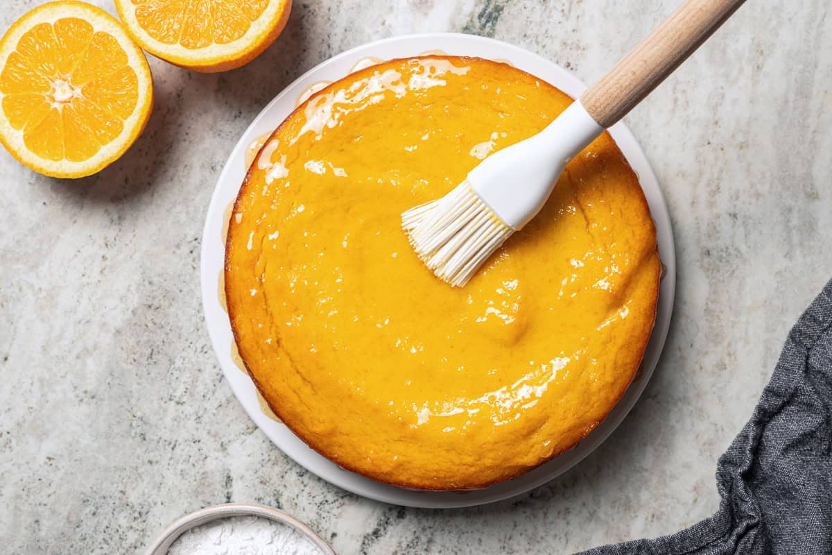 This orange cake recipe uses whole oranges, peels and all! It's moist and full of sweet citrus flavor, drenched in orange glaze. A pastry brush is brushing orange glaze over a cake.