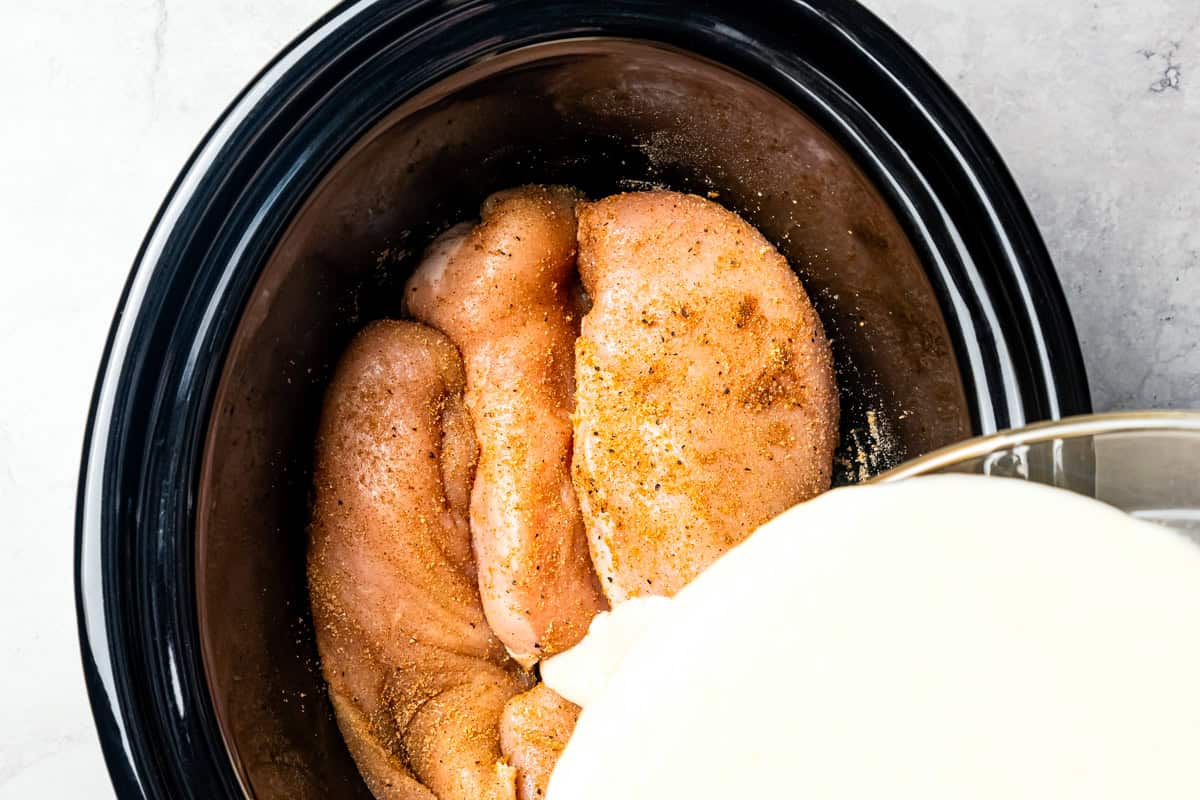 Pouring cream sauce over seasoned chicken in the slow cooker.