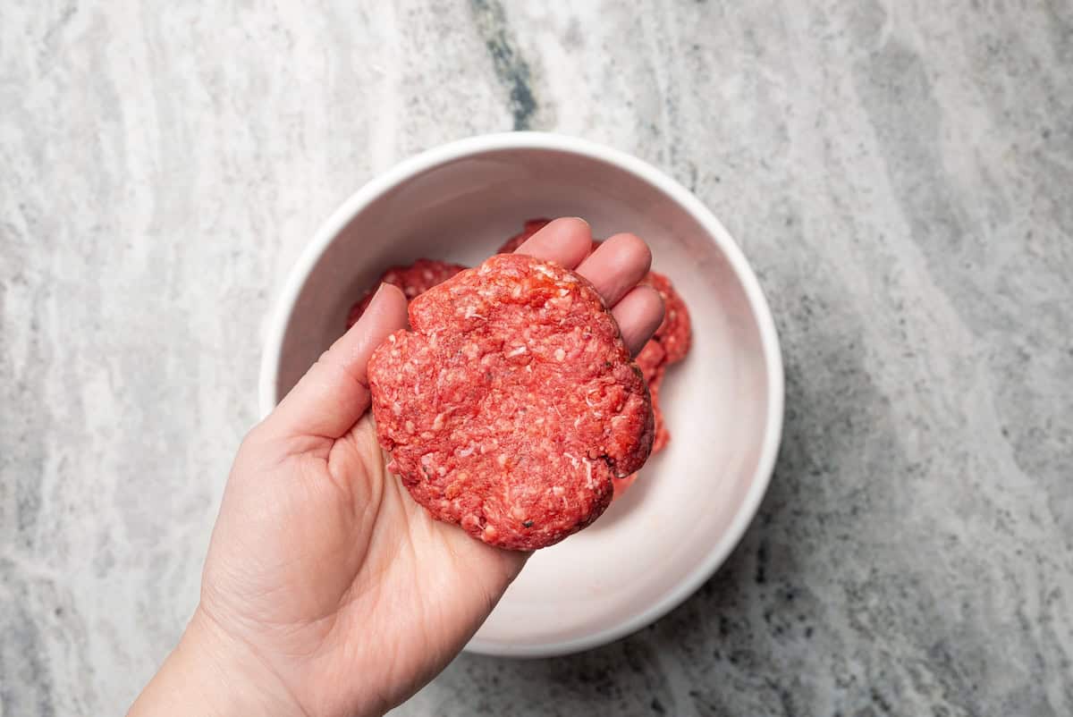 A hand holding a shaped hamburger patty over a bowl of hamburger meat.