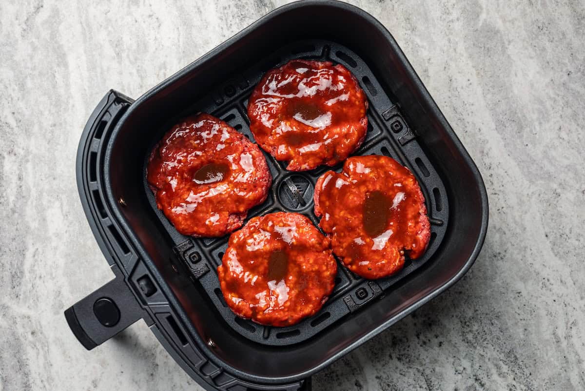 Four hamburger patties basted with BBQ sauce inside the air fryer basket.