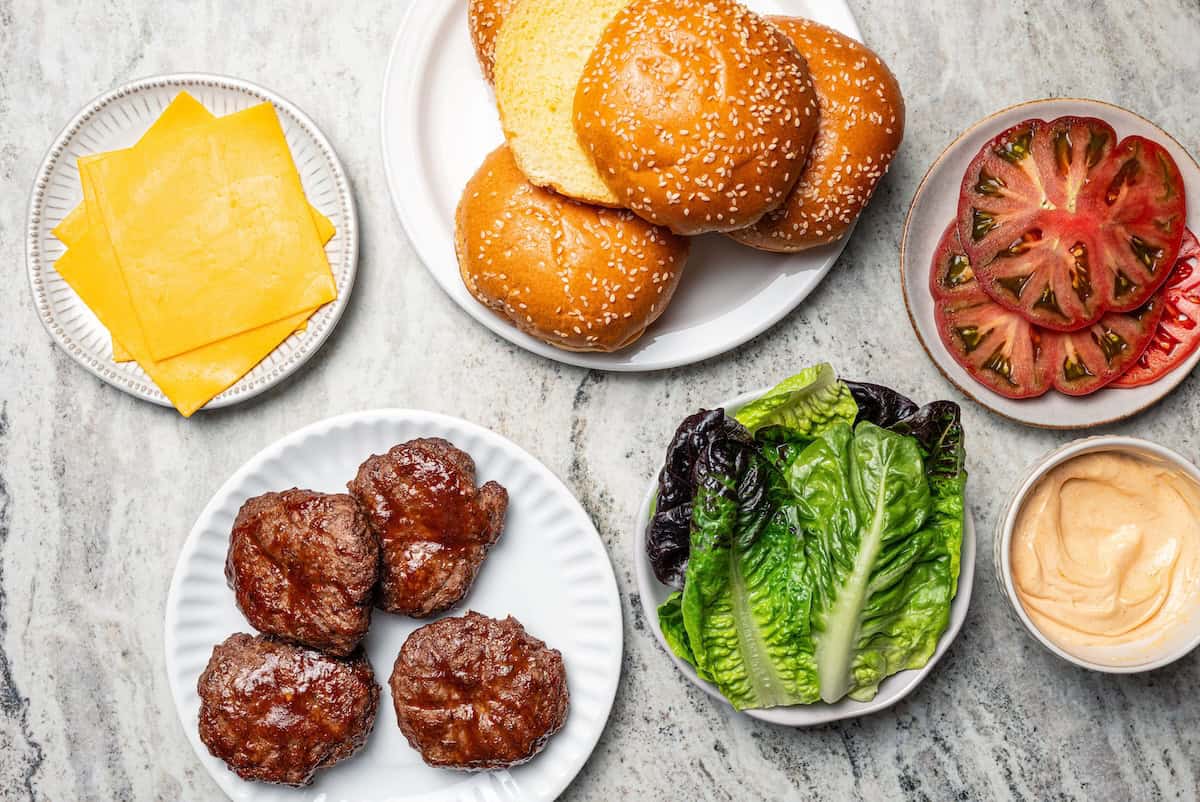 Overhead view of hamburger patties on a plate next to hamburger buns and garnishes.