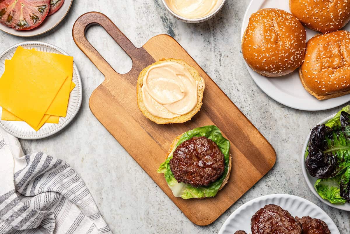Overhead view of a partially assembled hamburger next to a top bun dressed with spicy mayo on a cutting board.