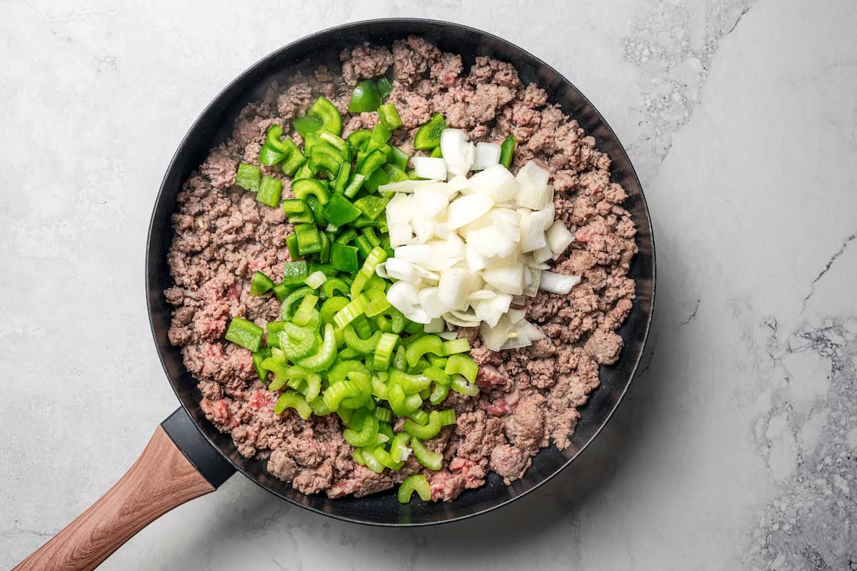 Diced onions, celery, and bell pepper added to browned ground meat in a skillet.