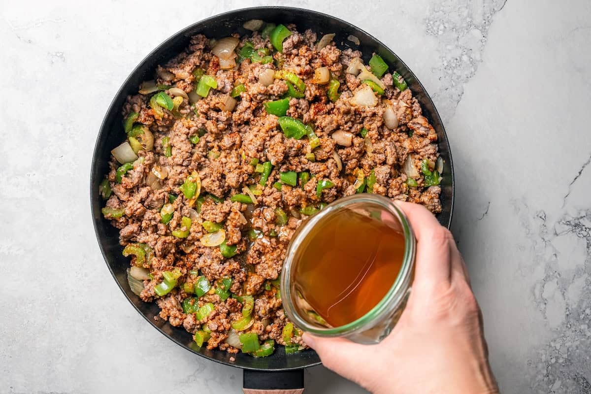 Pouring beef broth over the ground meat and veggies in a skillet.