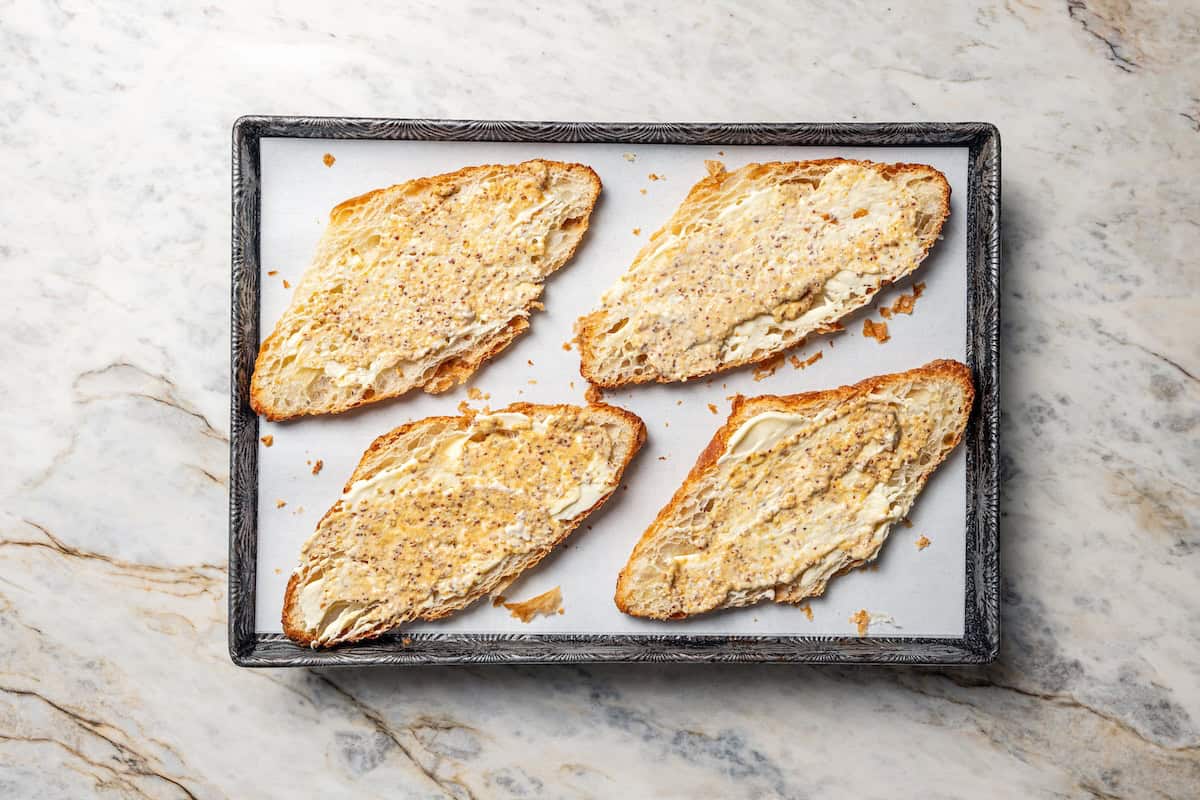 Four croissant halves spread with mayo and mustard on a baking sheet.