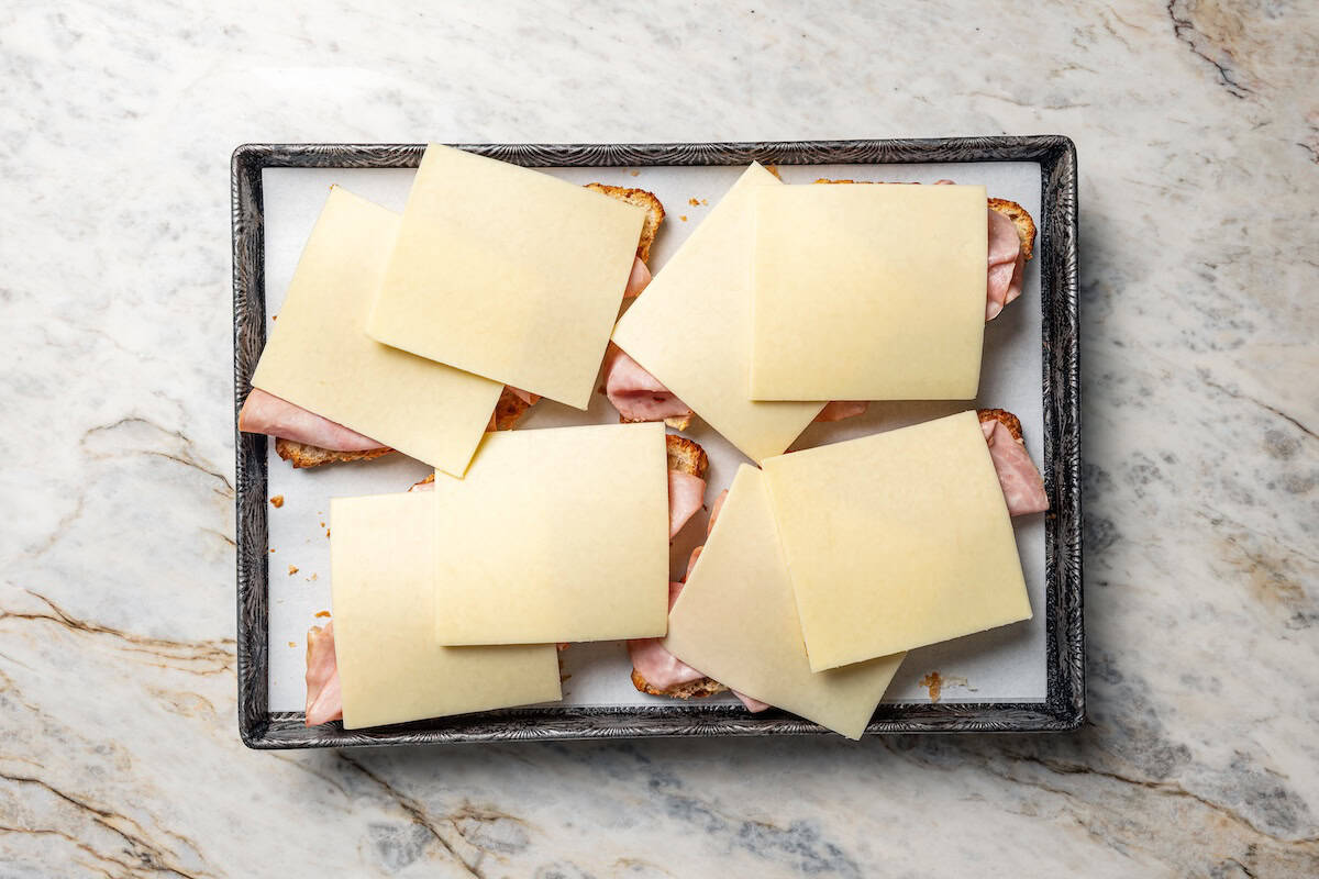 Four croissant halves topped with deli ham and cheese slices on a baking sheet.