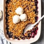 Homemade blueberry crisp in a baking dish, topped with three scoops of vanilla ice cream, with a spoon resting in the corner.