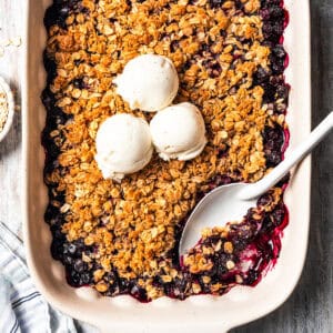 Homemade blueberry crisp in a baking dish, topped with three scoops of vanilla ice cream, with a spoon resting in the corner.