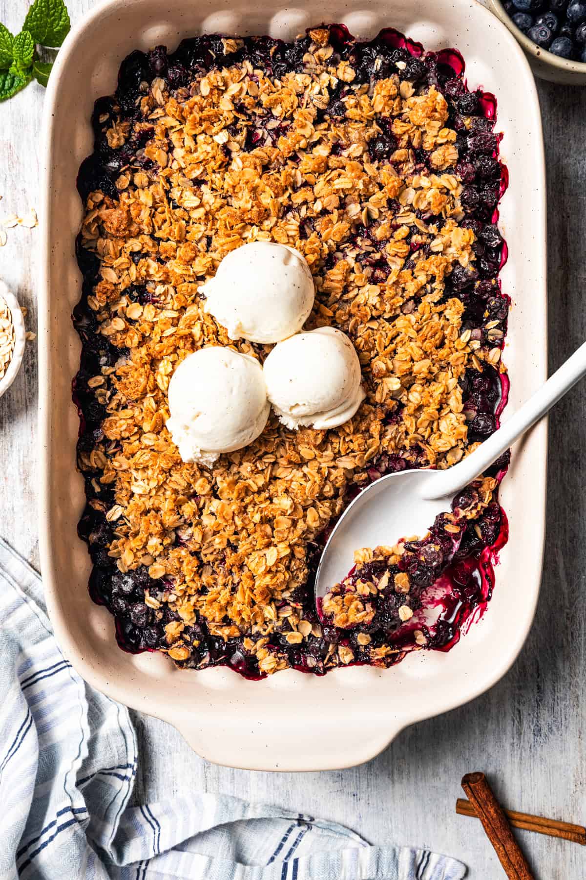Homemade blueberry crisp in a baking dish, topped with three scoops of vanilla ice cream, with a spoon resting in the corner.