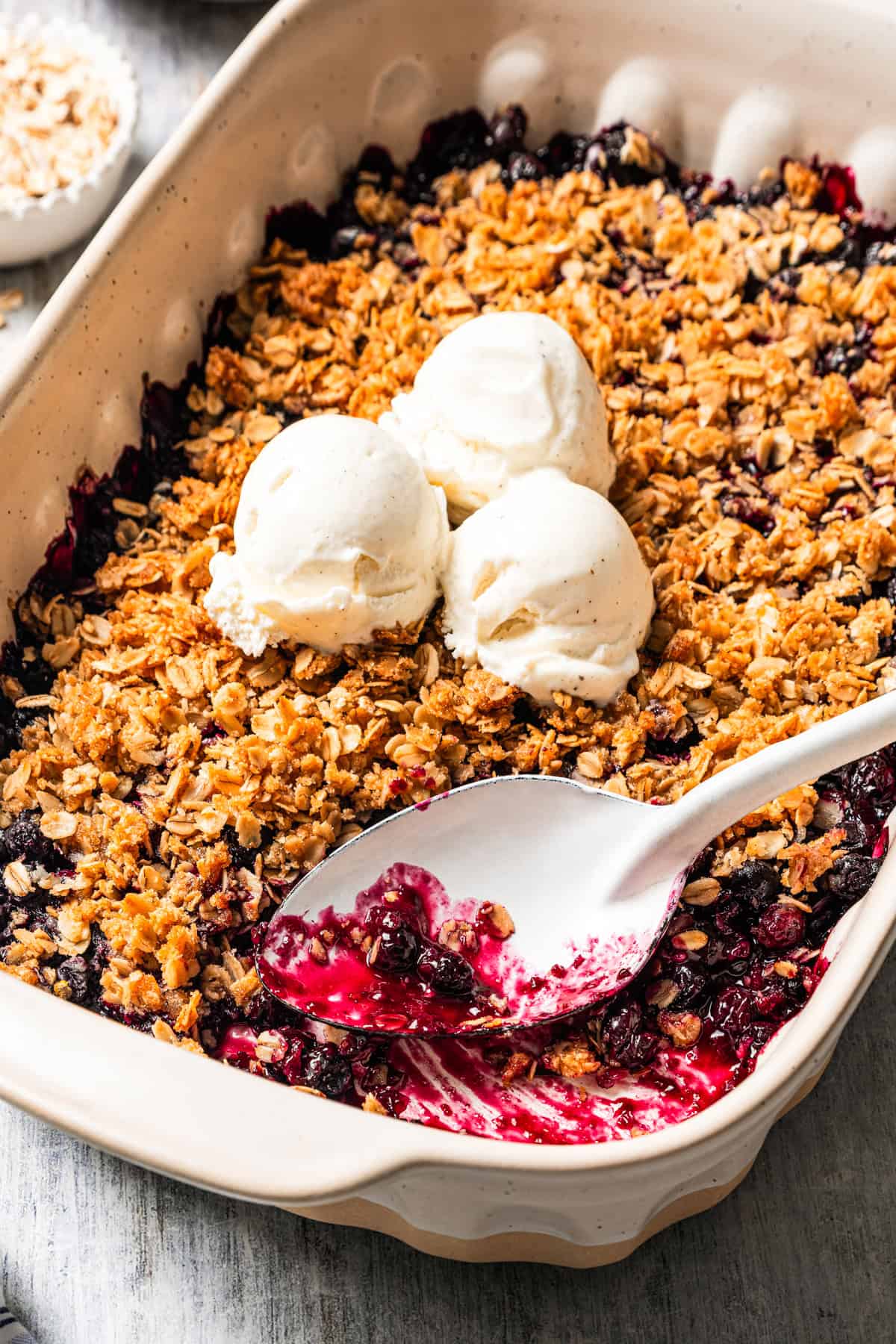 A blueberry crisp in a baking dish, topped with three scoops of vanilla ice cream, with a spoon resting in the corner.