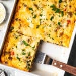 Overhead view of a breakfast lasagna in a baking dish, with a spatula resting in an open corner where a slice is missing.