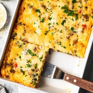 Overhead view of a breakfast lasagna in a baking dish, with a spatula resting in an open corner where a slice is missing.