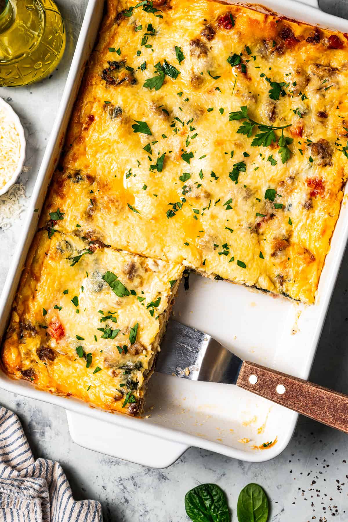 Overhead view of a breakfast lasagna in a baking dish, with a spatula resting in an open corner where a slice is missing.