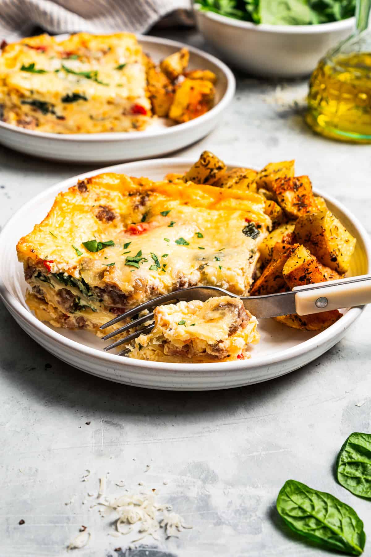 A fork cutting into a slice of breakfast lasagna next to breakfast potatoes on a plate.