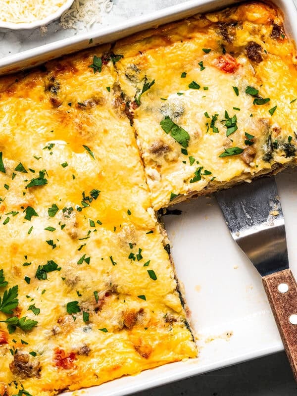 Breakfast lasagna in a casserole dish, with a spatula resting in an open corner where a slice is missing.