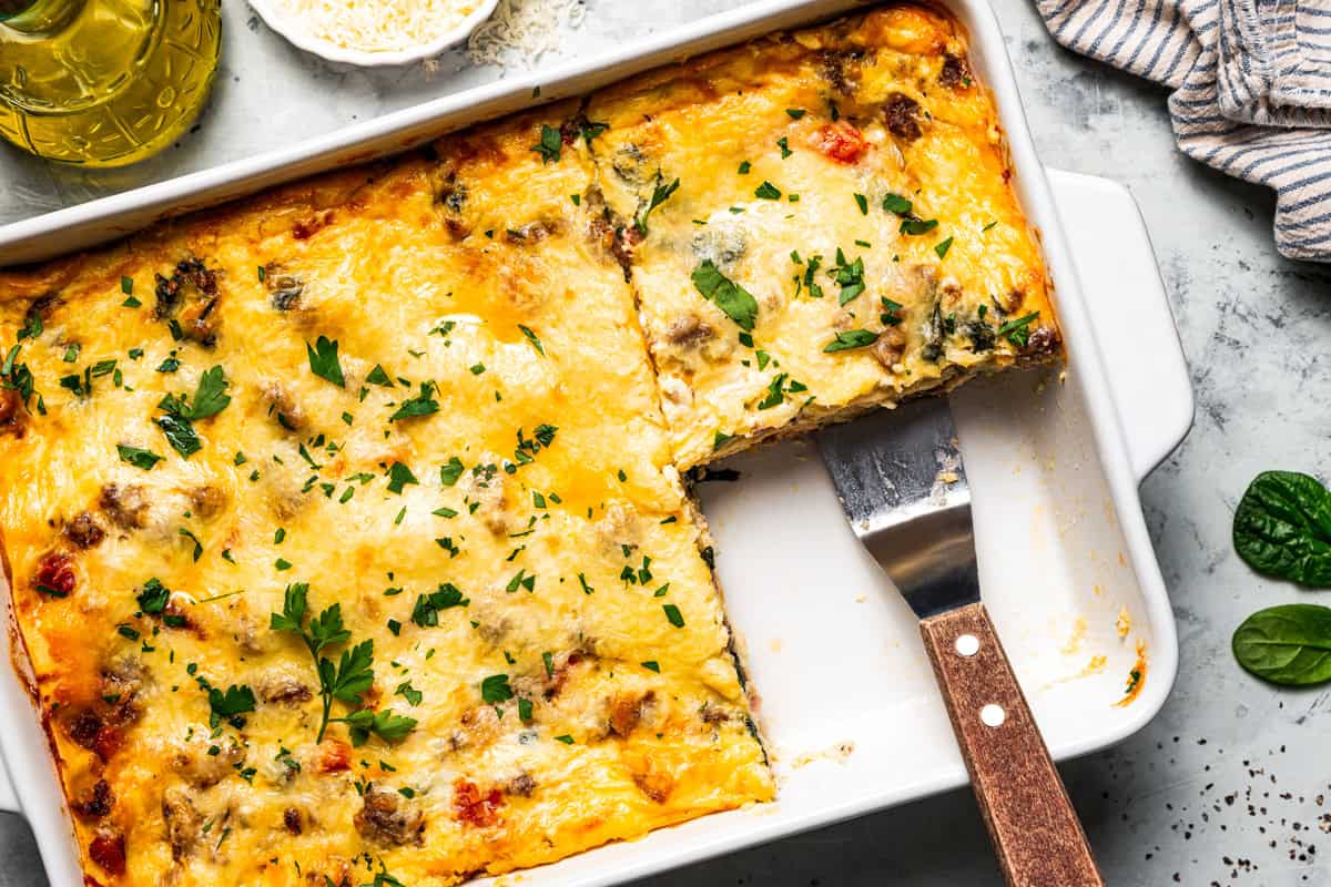 Breakfast lasagna in a casserole dish, with a spatula resting in an open corner where a slice is missing.