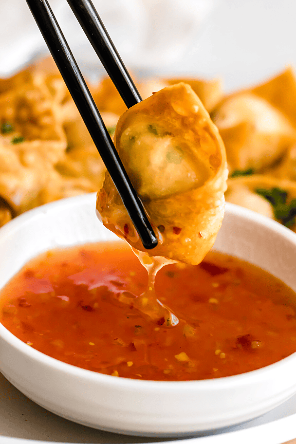 Chopsticks holding up a crab rangoon wonton over a bowl of sweet and sour sauce.