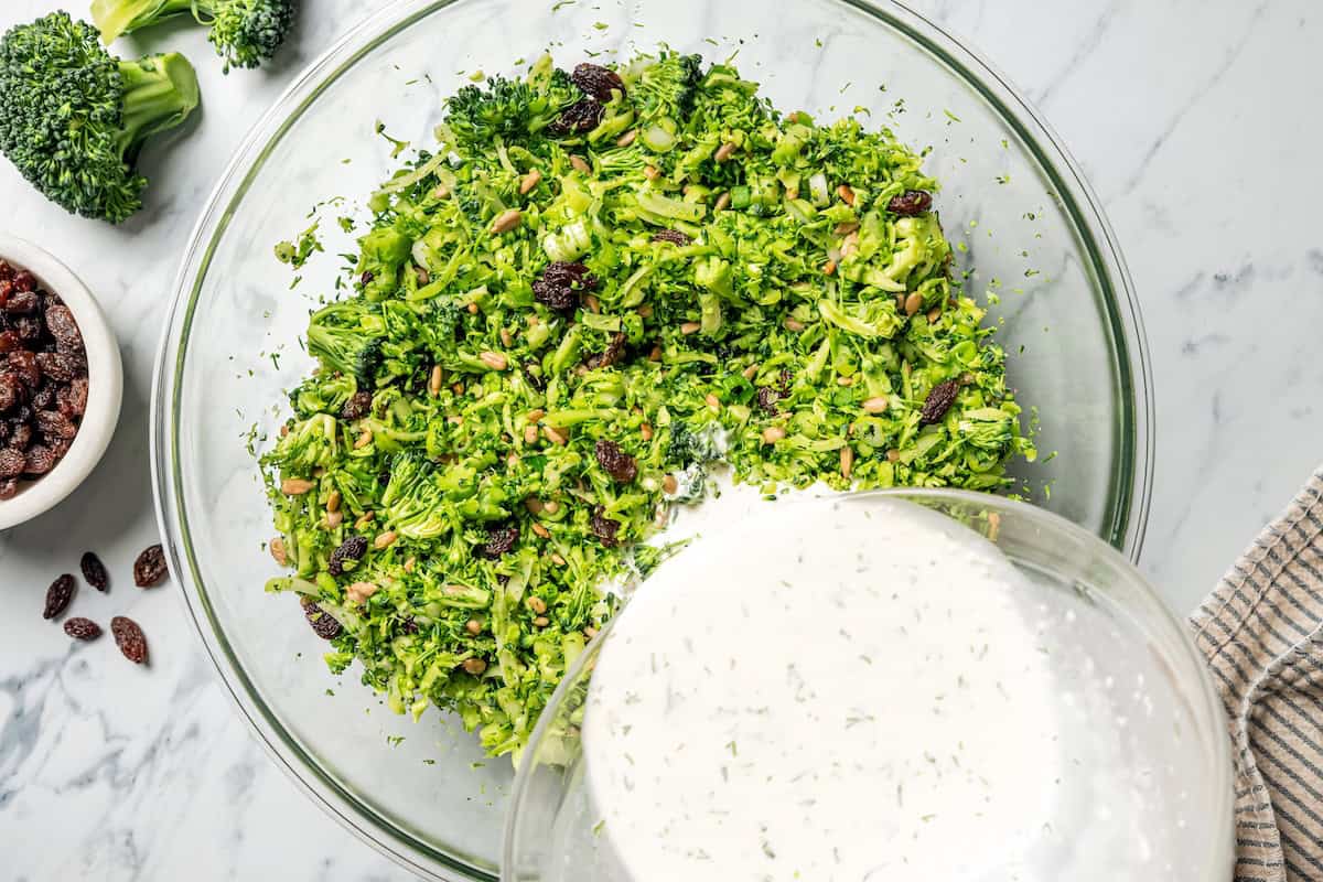 This broccoli slaw recipe is a fresh, 10-minute side dish with crunchy broccoli, sunflower seeds, and raisins in creamy buttermilk dressing. Pouring dressing over a bowl of broccoli slaw ingredients.
