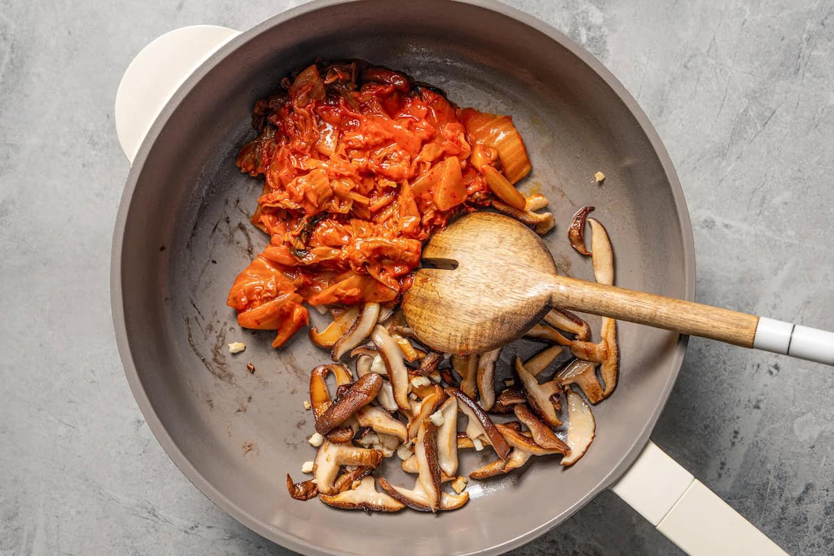 This quick and easy kimchi ramen recipe packs a spicy, savory punch with instant noodles, kimchi, shiitake mushrooms, and gochujang broth. Kimchi and mushrooms sautéing in a skillet.