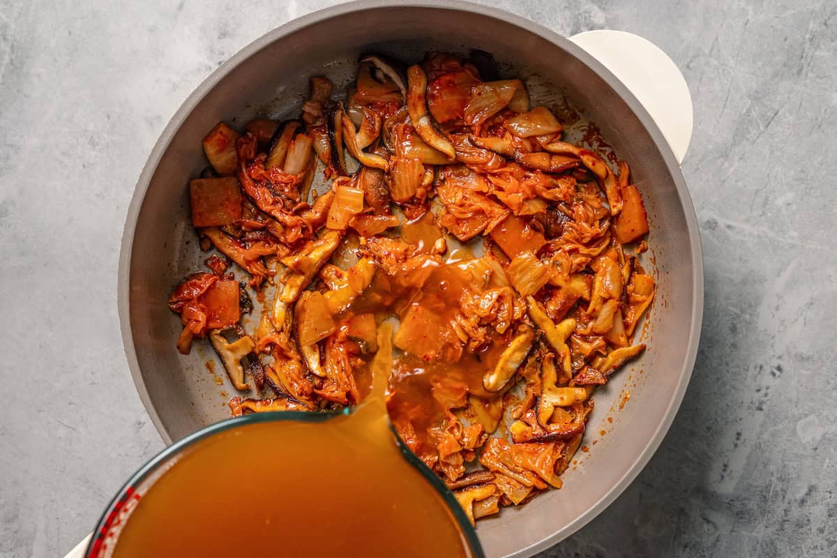 This quick and easy kimchi ramen recipe packs a spicy, savory punch with instant noodles, kimchi, shiitake mushrooms, and gochujang broth. Pouring liquid into a skillet with kimchi and mushrooms.