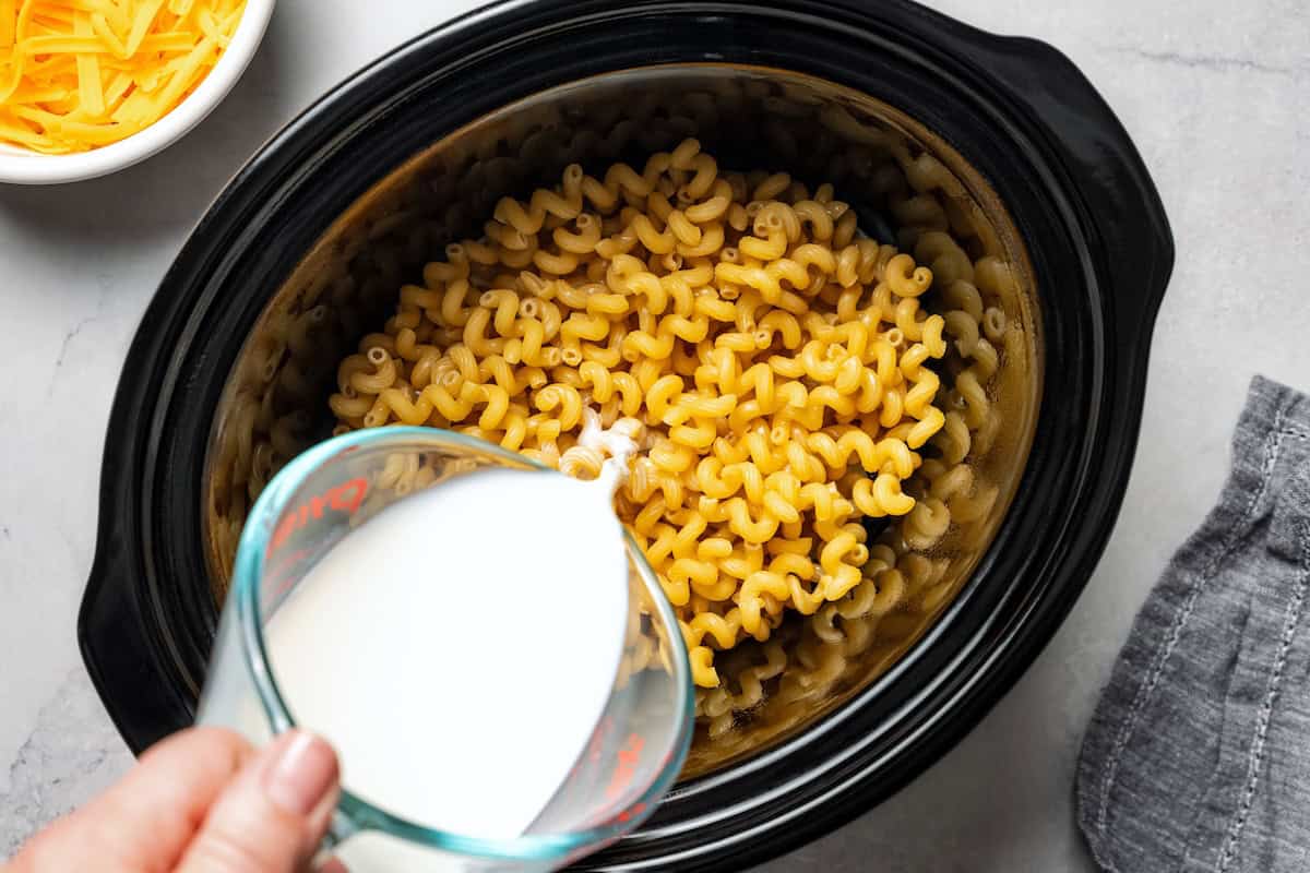 Ultra-creamy slow cooker mac and cheese is a dump-and-go version of my homemade mac and cheese recipe! Perfect for potlucks and holidays. Pouring milk over rinsed pasta inside the slow cooker.