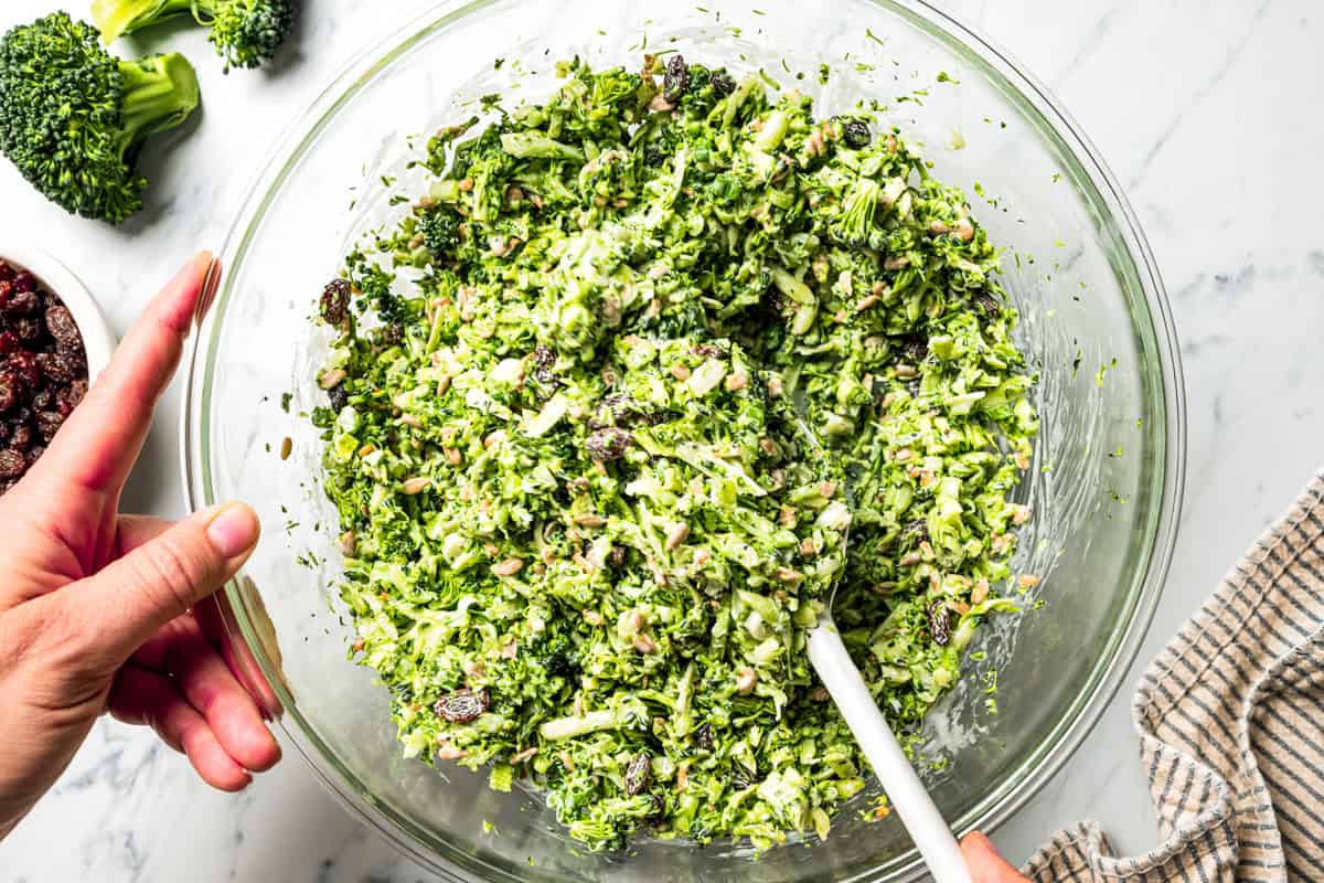 This broccoli slaw recipe is a fresh, 10-minute side dish with crunchy broccoli, sunflower seeds, and raisins in creamy buttermilk dressing. A hand holding a bowl of broccoli slaw while mixing together the ingredients.