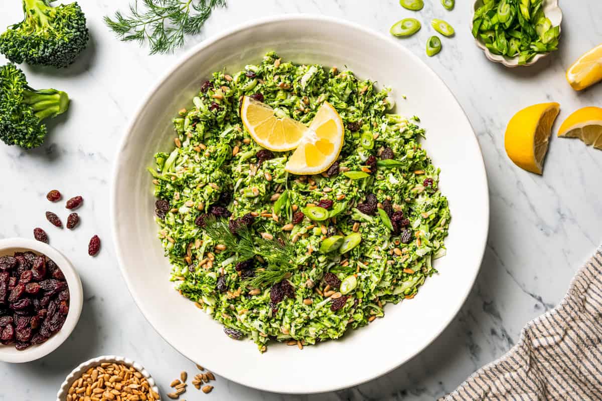 This broccoli slaw recipe is a fresh, 10-minute side dish with crunchy broccoli, sunflower seeds, and raisins in creamy buttermilk dressing. Broccoli slaw served in a salad bowl and garnished with lemon wedges and fresh green herbs.