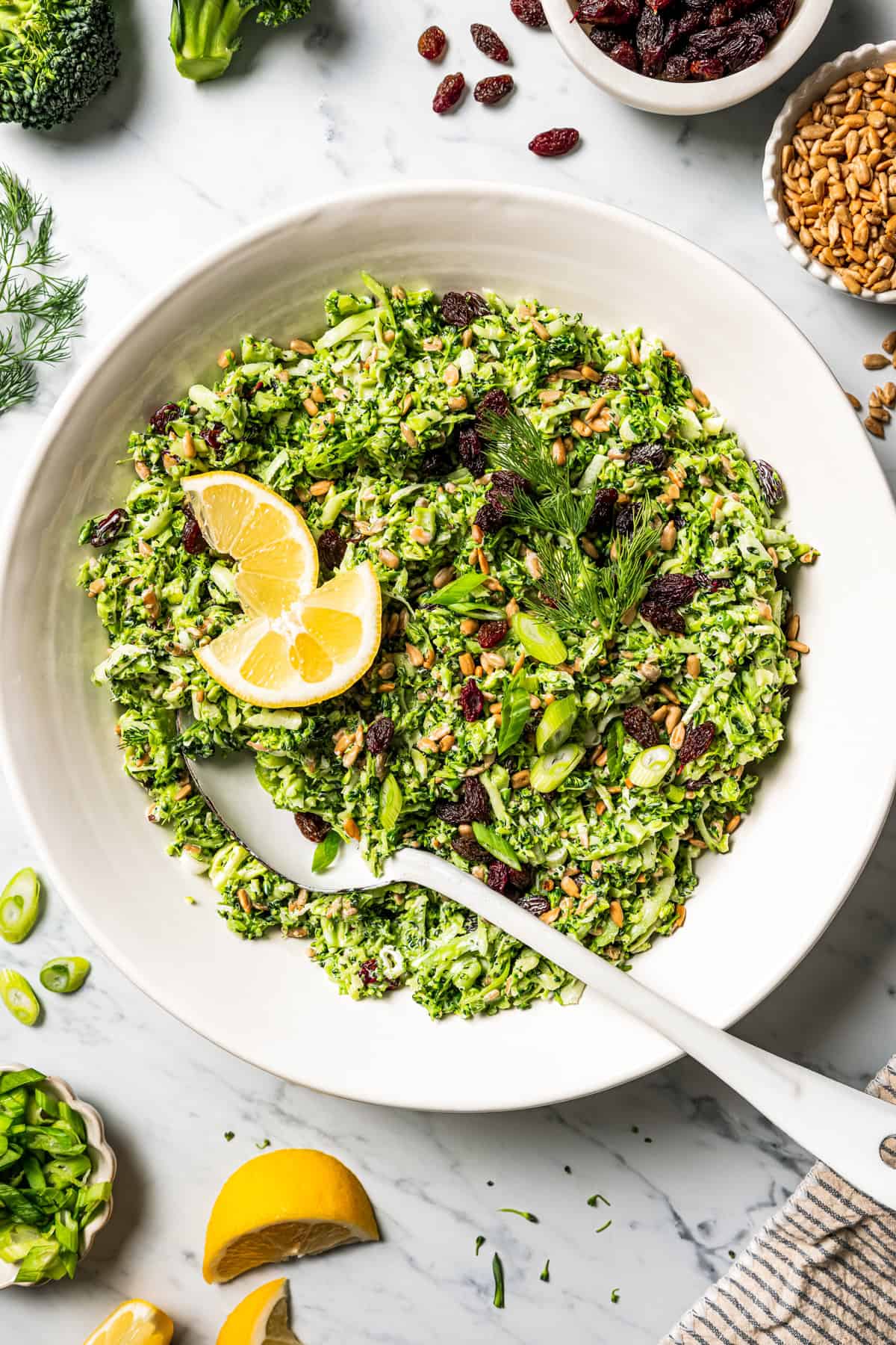 This broccoli slaw recipe is a fresh, 10-minute side dish with crunchy broccoli, sunflower seeds, and raisins in creamy buttermilk dressing. A bowl of broccoli slaw garnished with lemon wedges.